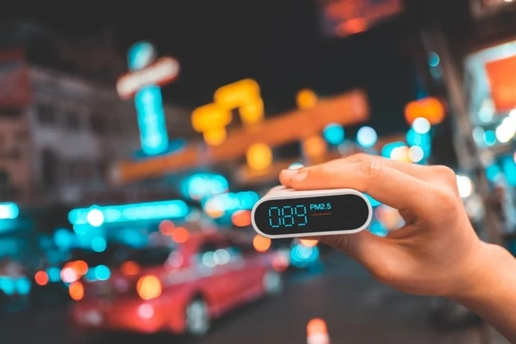 Hand holding an air quality monitoring device, with a blurry city in the background, with lots of bright lights.