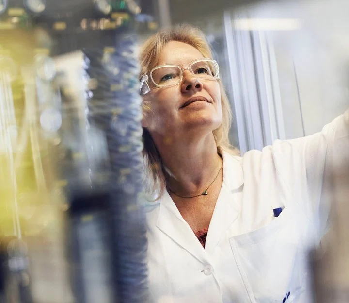 A woman wearing a lab coat and safety specs looks at a chemical set up.