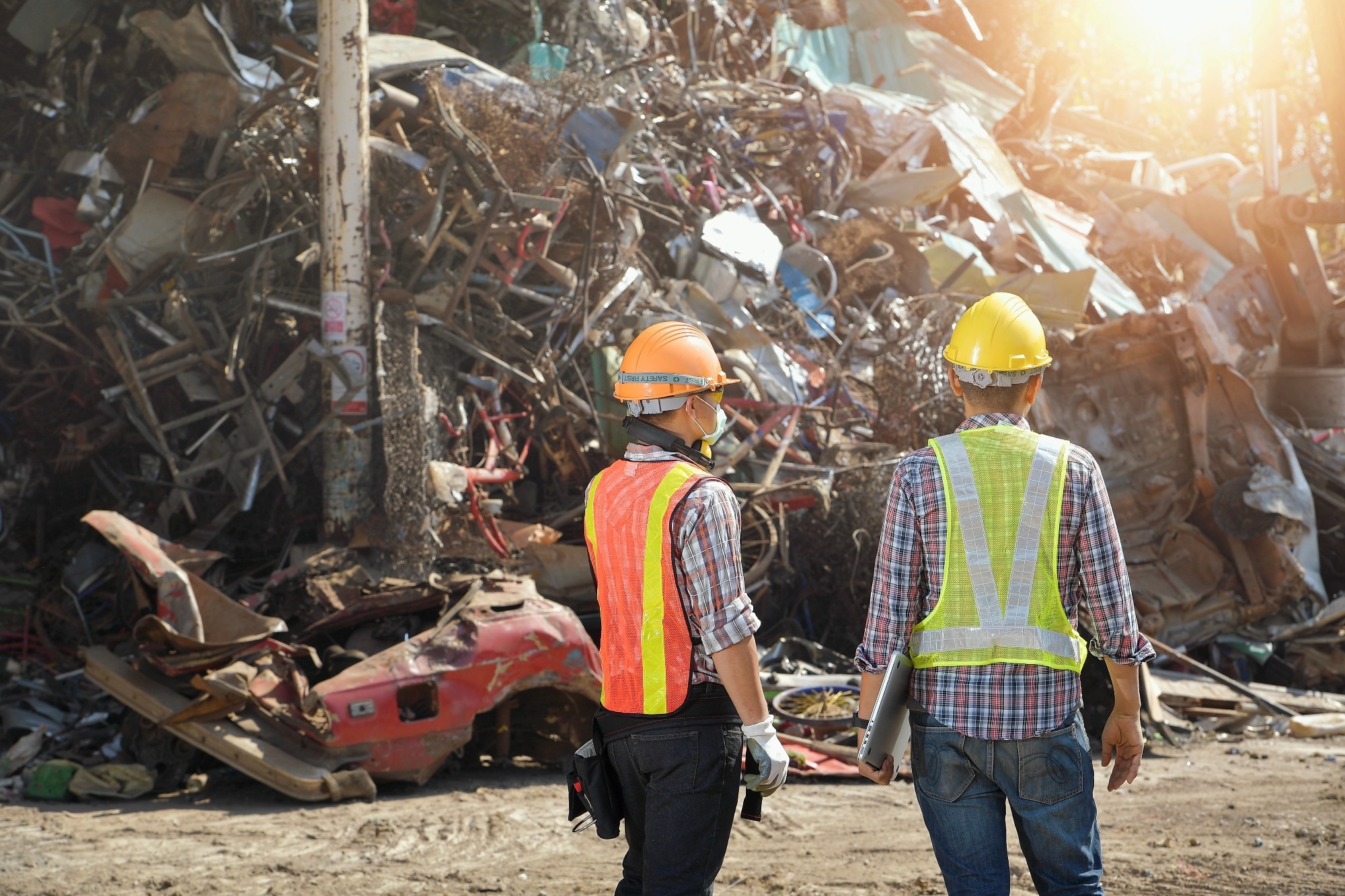 Two workers in safety gear, including hard hats and reflective vests, are standing in front of a large pile of scrap metal and debris at a junkyard or recycling facility. One worker holds a laptop, while the other has gloves and is observing the scene.